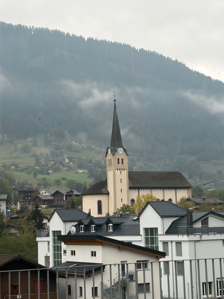 Church spire above Fiesch rooftops against a forested mountainside.