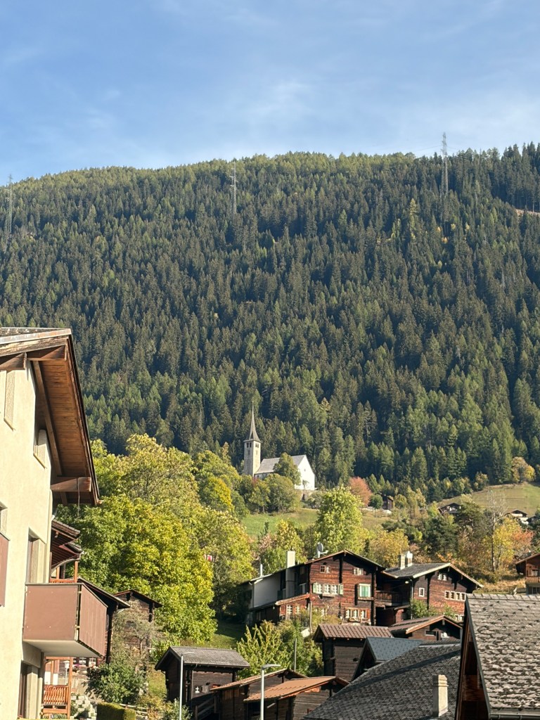 Fiesch rooftops and a white church below a green and gold hillside.