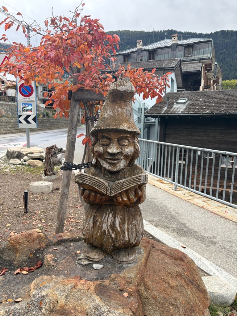 Wooden gnome holding an open book beneath red autumn leaves.