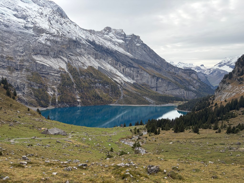 Turquoise Lake Oeschinen below snow-dusted Alpine peaks, with autumn trees, grassy slopes, and a mountain hut in the foreground.