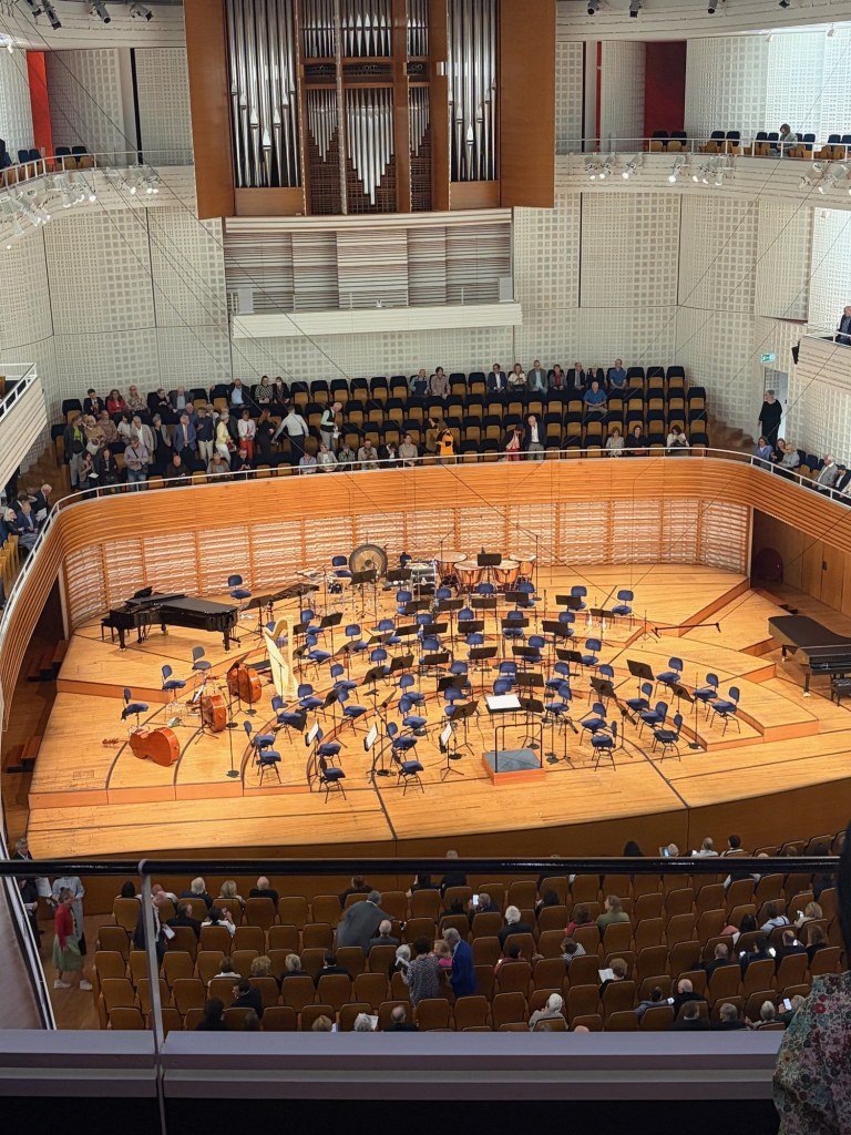 An orchestra stage with a grand piano, harp, and rows of chairs beneath a pipe organ in Lucerne’s concert hall.
