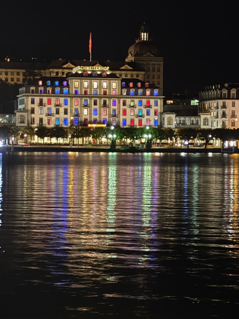 Colorful illuminated windows of the Schweizerhof reflect across dark lake water.