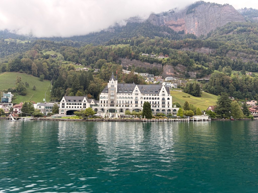 A grand white waterfront building stands between teal lake water and a misty, wooded mountainside near Vitznau.