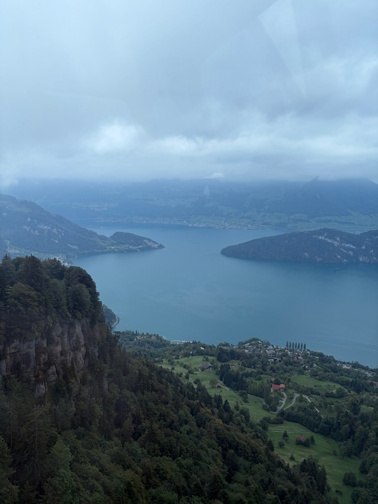 Lake Lucerne stretches between wooded headlands beneath low clouds, seen from above green fields and cliffs.