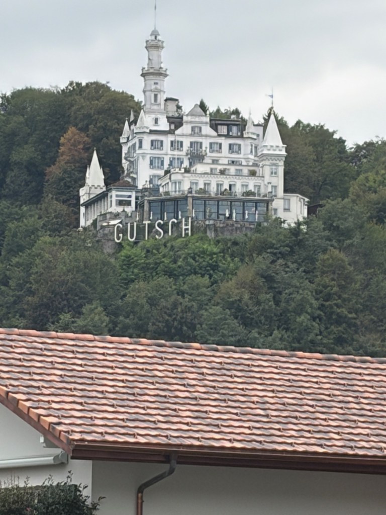 A white turreted building and large Gütsch sign rise above a wooded hillside, with a red-tiled roof in the foreground.