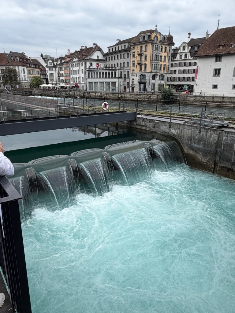 Turquoise water spills over a weir below historic buildings along the Reuss.