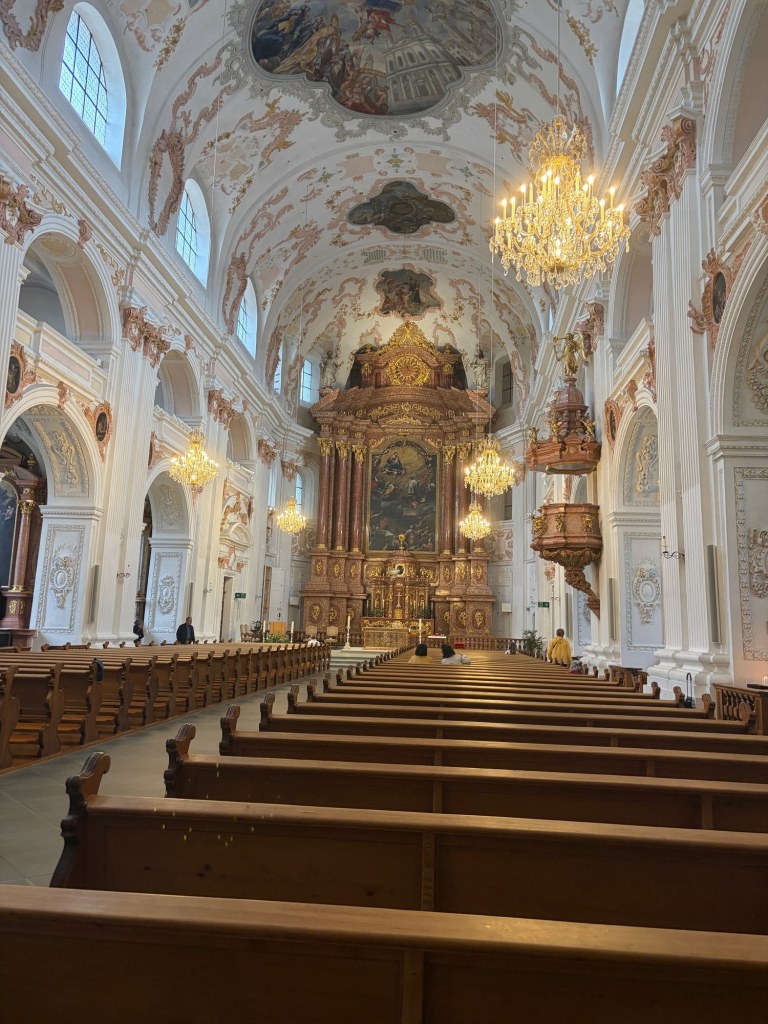 Painted ceilings, white columns, golden chandeliers, and wooden pews lead toward the Jesuit Church altar.