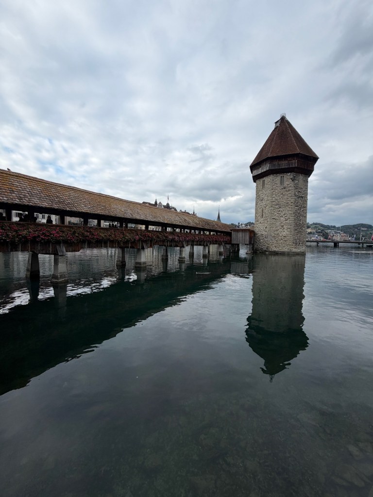 Chapel Bridge and its stone water tower reflected in the calm Reuss beneath a cloudy sky.