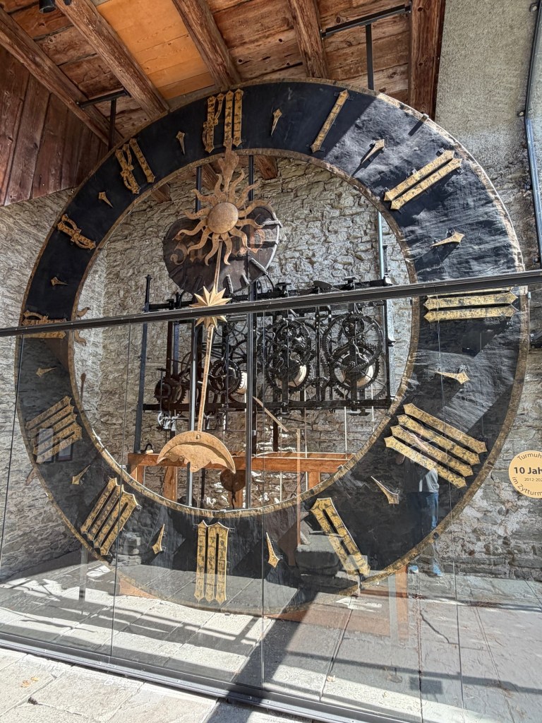 A large historic clock face with gold numerals and exposed mechanisms behind protective glass.