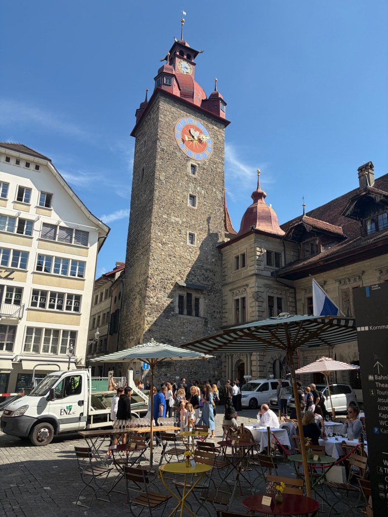 A stone clock tower rises above café tables and people in a sunny Lucerne square.