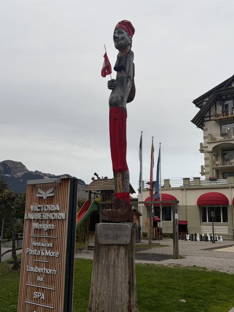 Tall carved wooden figure in red trousers holding a Swiss flag in Wengen.