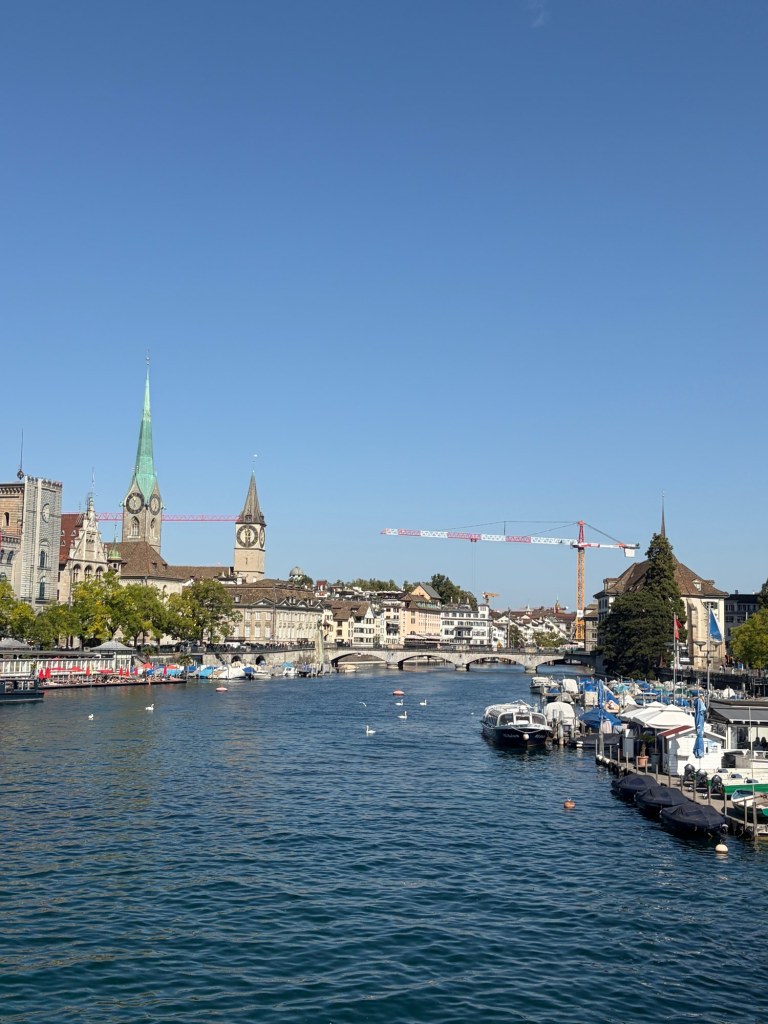 Blue river water and moored boats beneath Zürich’s skyline, with two church towers and a bridge.