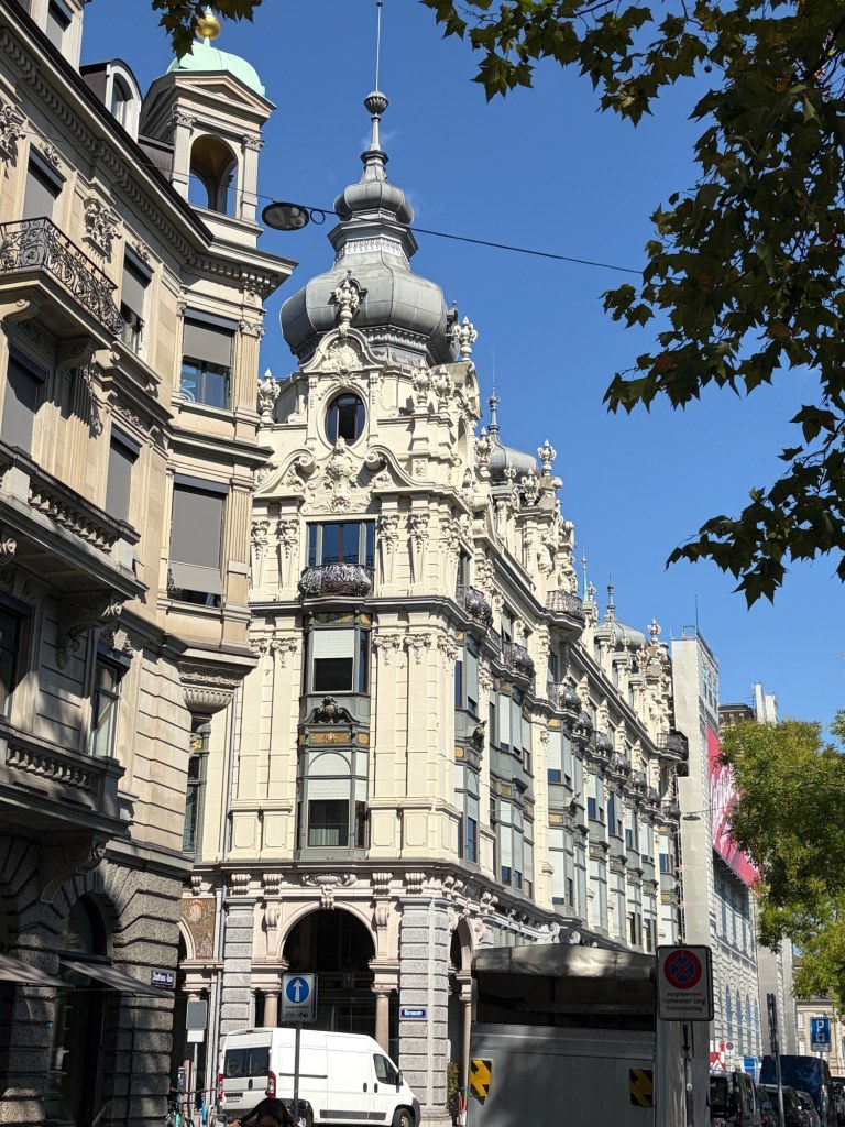 An ornate pale stone building with sculpted figures and a domed corner tower, framed by leaves.