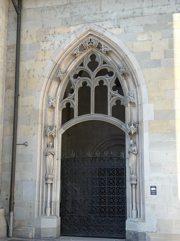 A pointed stone arch with intricate tracery and two carved figures surrounds a dark iron gate.