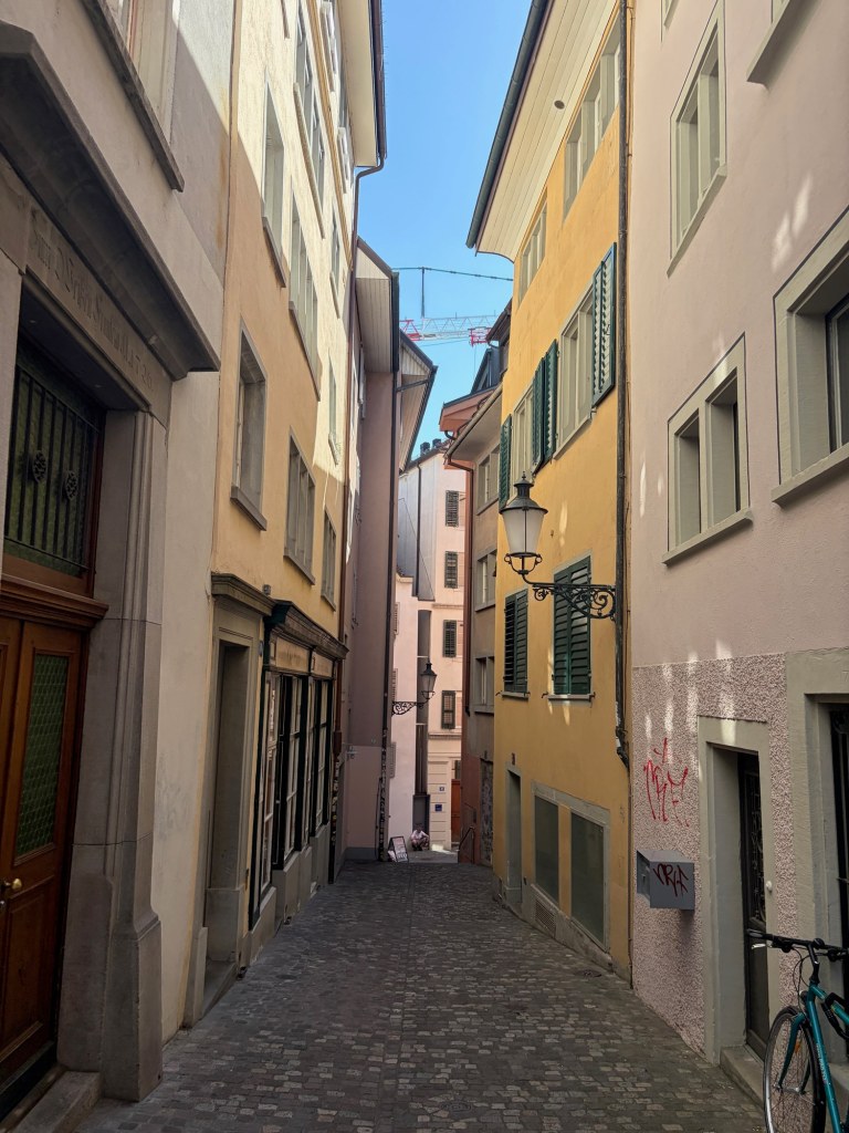 A narrow cobblestone lane between pastel buildings with green shutters in Zürich.