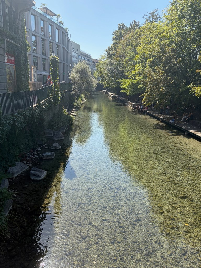 Clear water in a narrow Zürich canal reflects leafy trees beside buildings and waterside walkways.